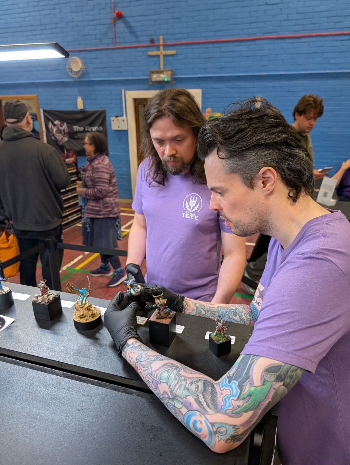 Two Iron Thistle judges in purple show t-shirts examining a painted miniature